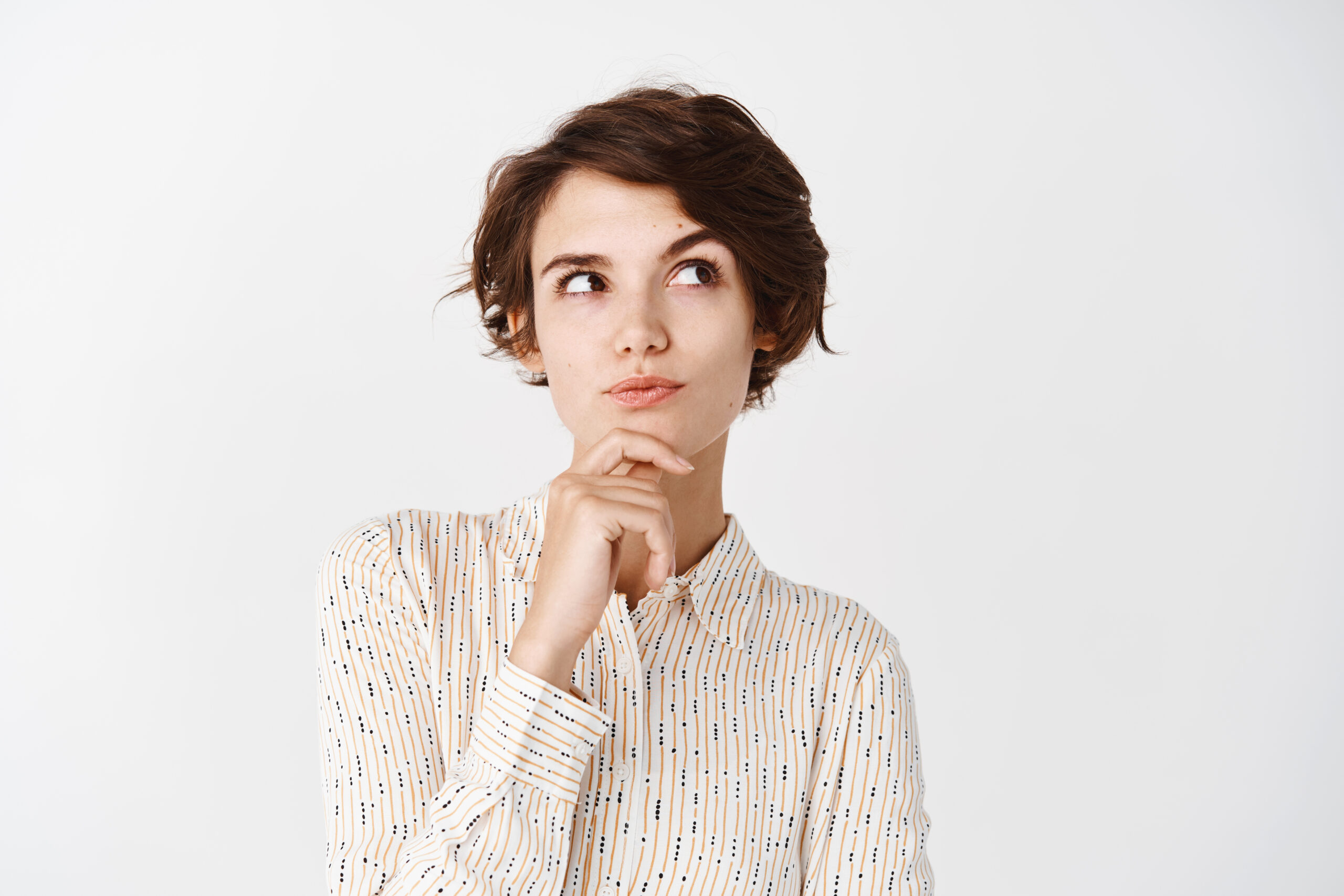 Pensive girl in blouse touching chin, looking at upper right corner and thinking, making choice, standing over white background. Copy space